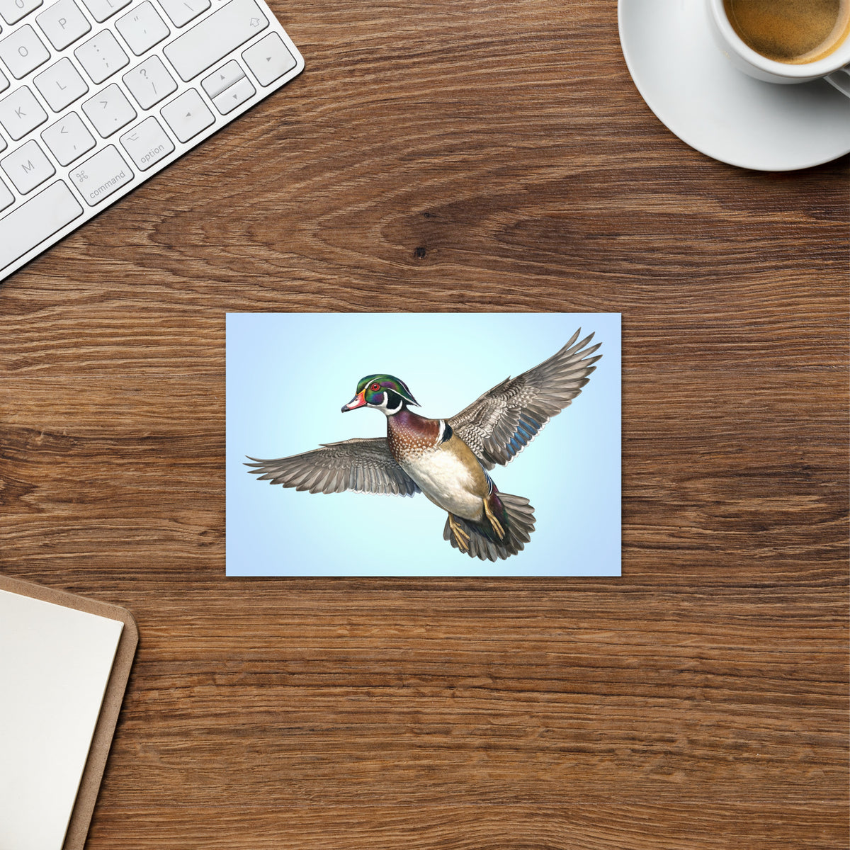 Product mockup of the Wood Duck Greeting Card, shown on a desk with a coffee cup, keyboard and clipboard.